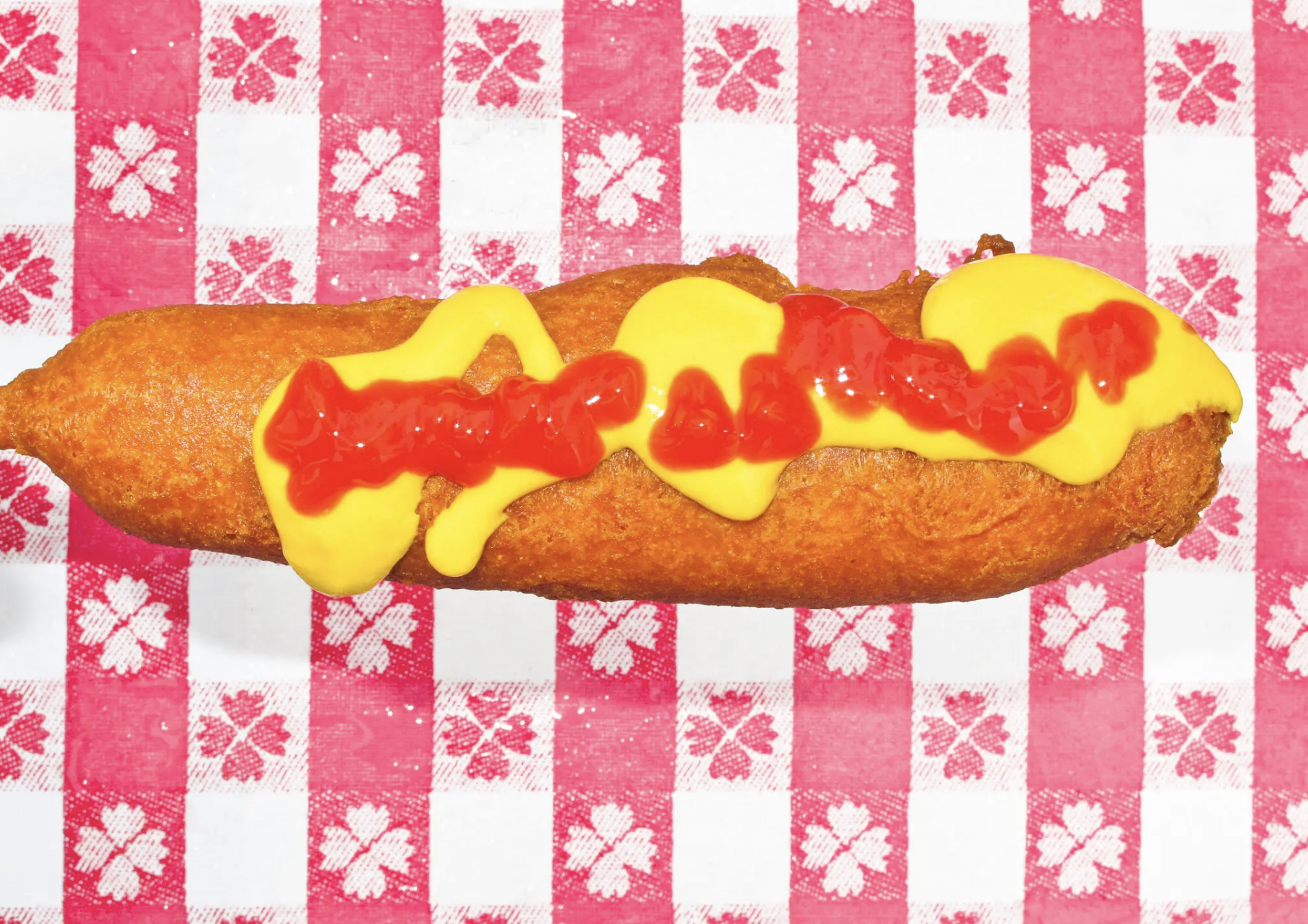 a corndog with mustard and ketchup in front of a garish red and white patterned tablecloth